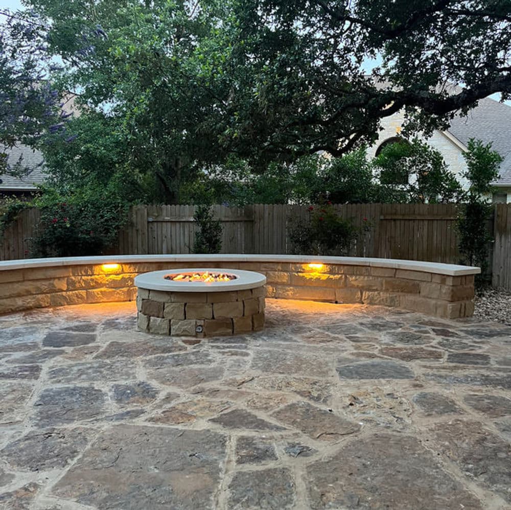 Outdoor stone patio with a circular fire pit and seating area, surrounded by lush greenery.