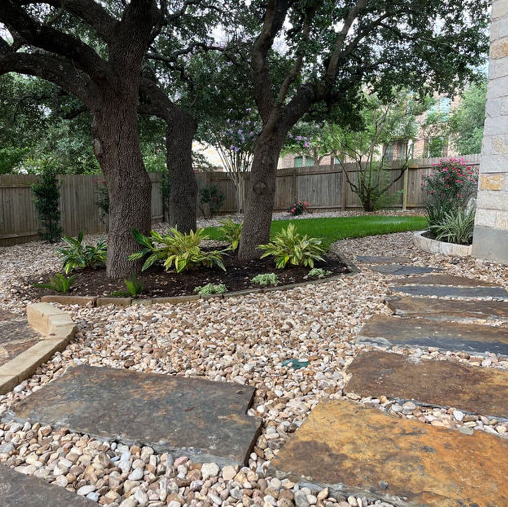 Backyard landscape with stone pathway, lush plants, and mature trees in a serene setting.