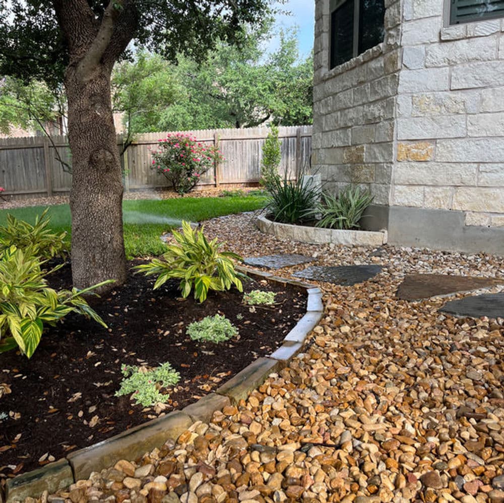 Landscaped yard featuring stone pathways, greenery, and a tree near a house.