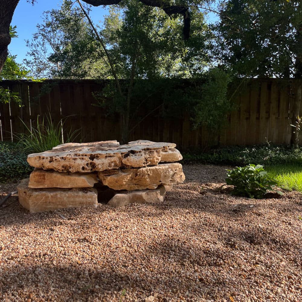 Natural stone bench on gravel in a serene backyard landscape with trees and greenery.