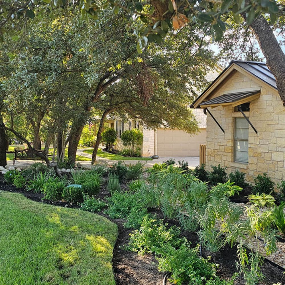 Lush garden with vibrant plants beside a charming stone house in a sunny landscape.